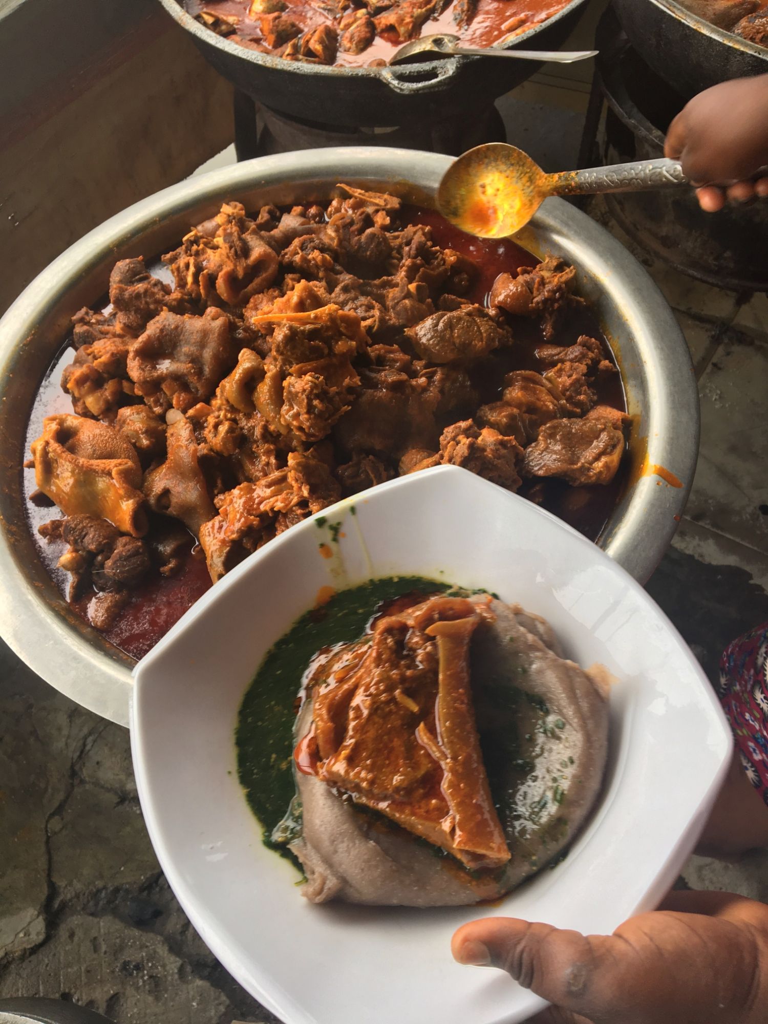 A plate of amala with ewedu and ponmo in stew, held beside a buka pot of assorted meat