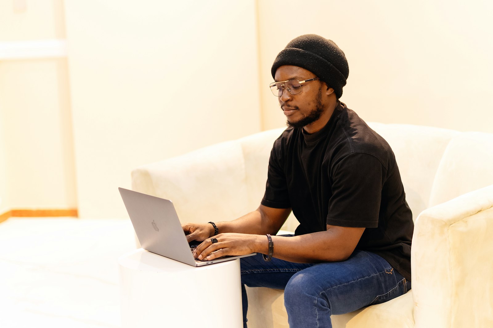 A young Black man in a black T-shirt and beanie typing on a laptop on a cream sofa