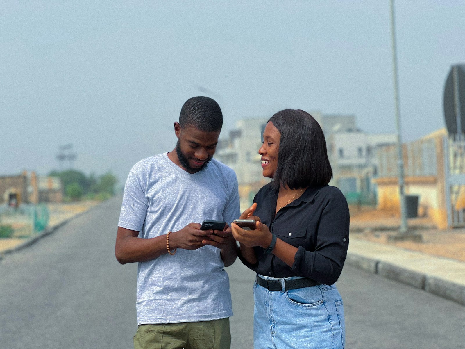 A young Black man and woman standing on a quiet street, each holding a smartphone, smiling as they compare their screens
