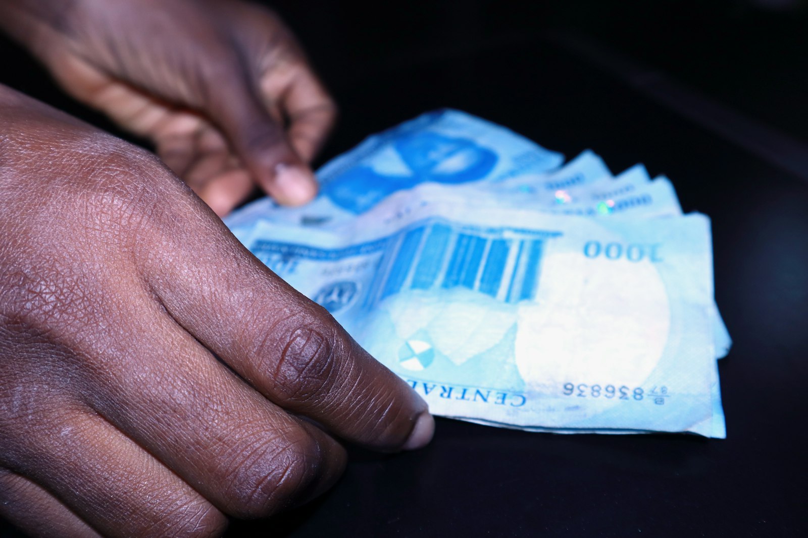 Close-up of two dark-skinned hands fanning out a few blue ₦1,000 notes on a dark table