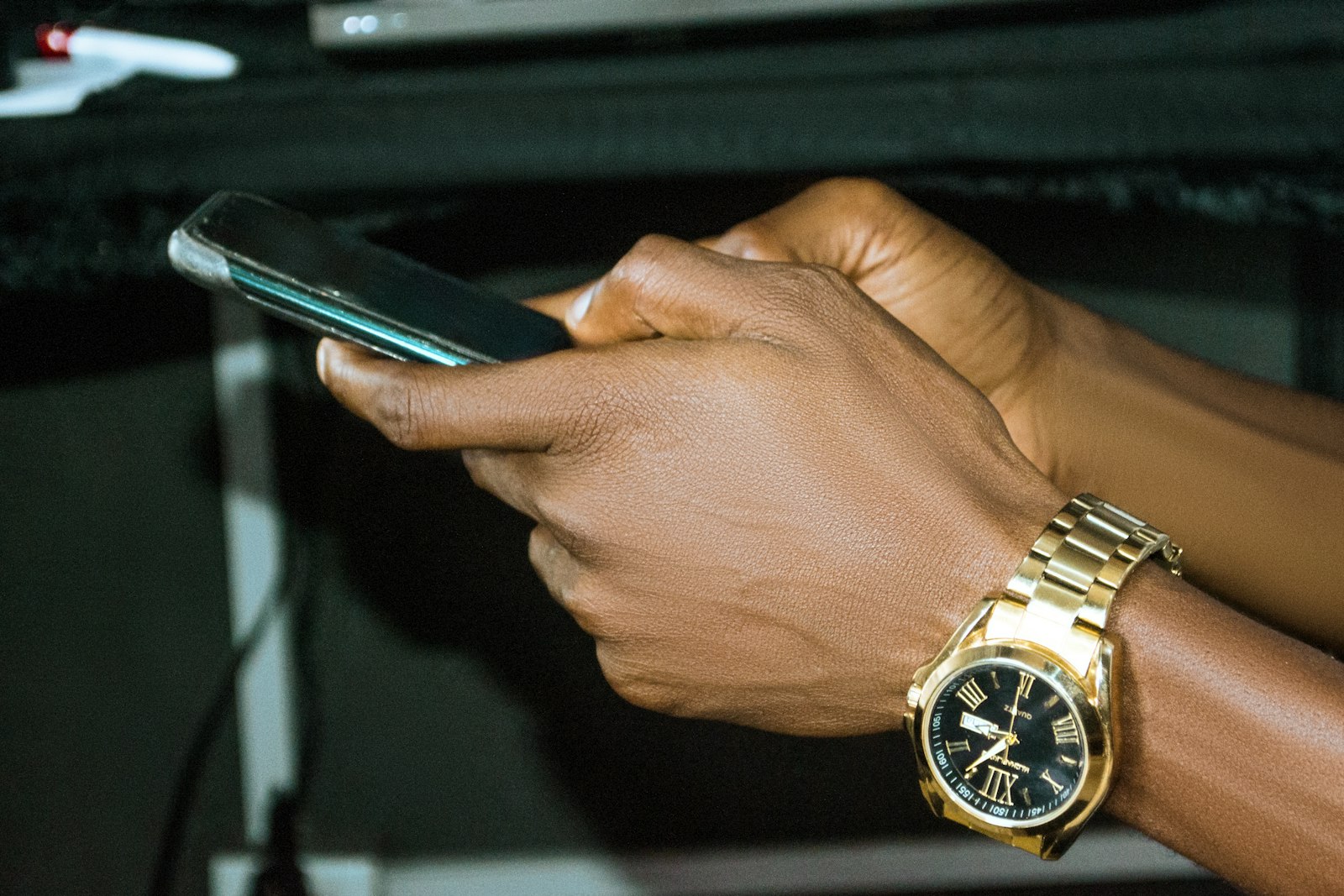 Close-up of a man's dark-skinned hands typing on a smartphone, a gold wristwatch on his wrist