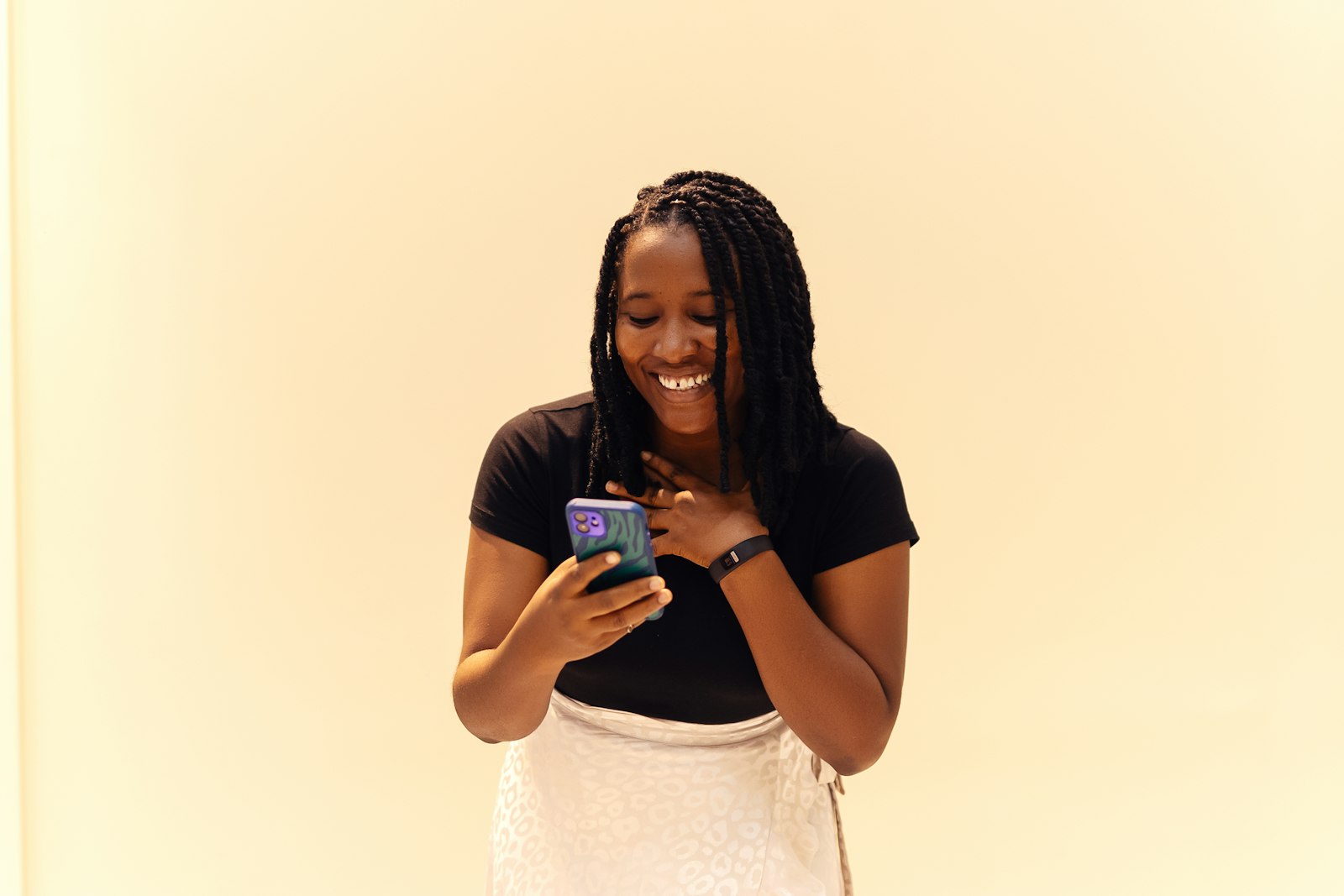 A woman with braids smiling at her phone with one hand on her chest, standing against a plain cream wall