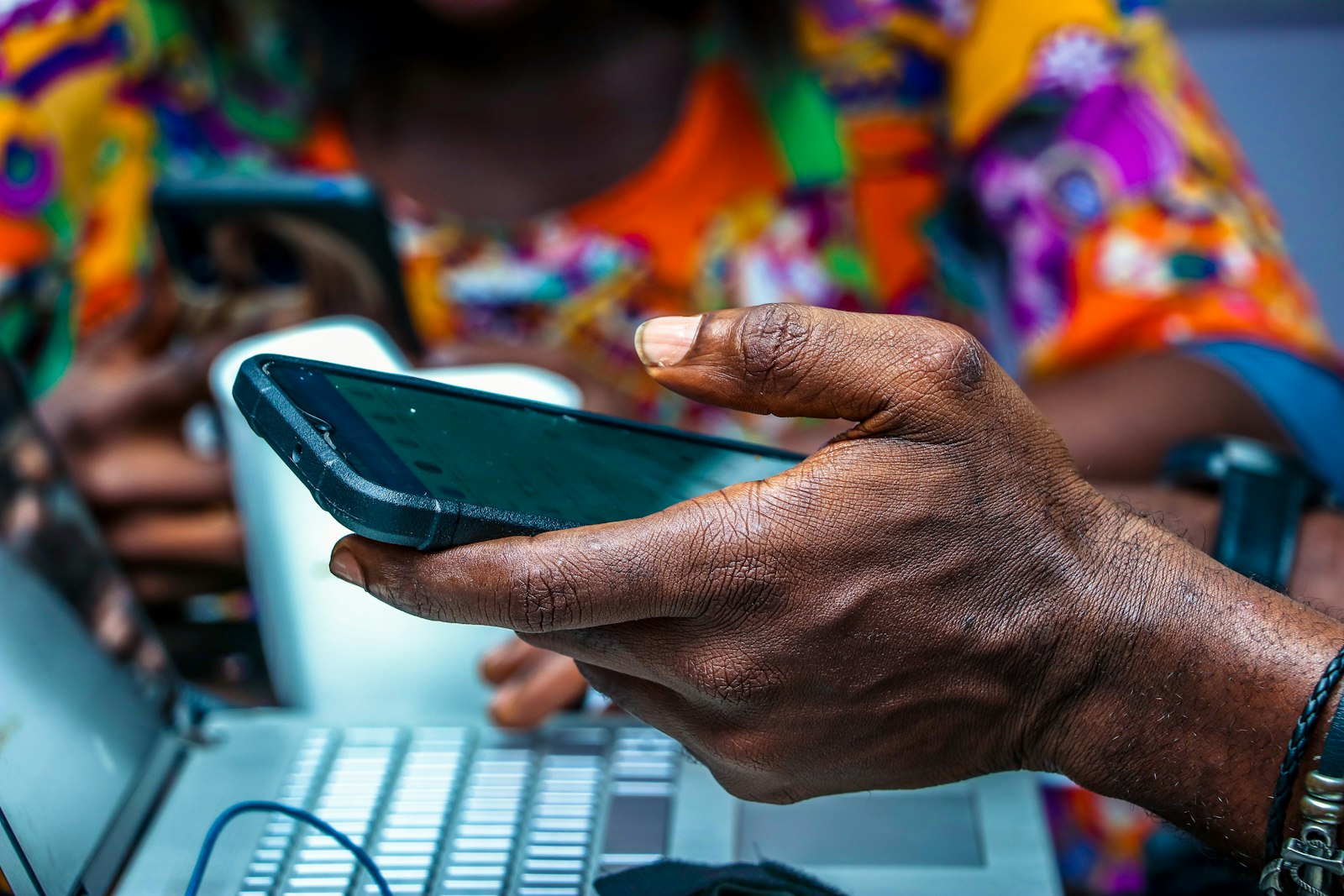 A hand holding a phone over an open laptop, checking the screen mid-work, with a colourful print shirt out of focus behind