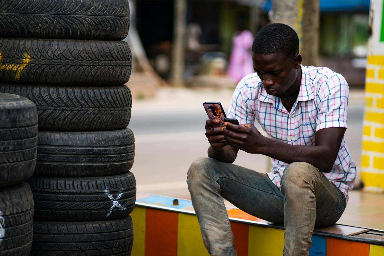 A man sitting beside a stack of tyres at a roadside shop, comparing two phone screens in his hands